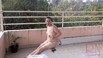 女性は傘の下で雨から隠れて、ぶら下がっているブランコで揺れています。 庭の夏の雨。 テラスに透明な傘をさした裸のヌーディスト。